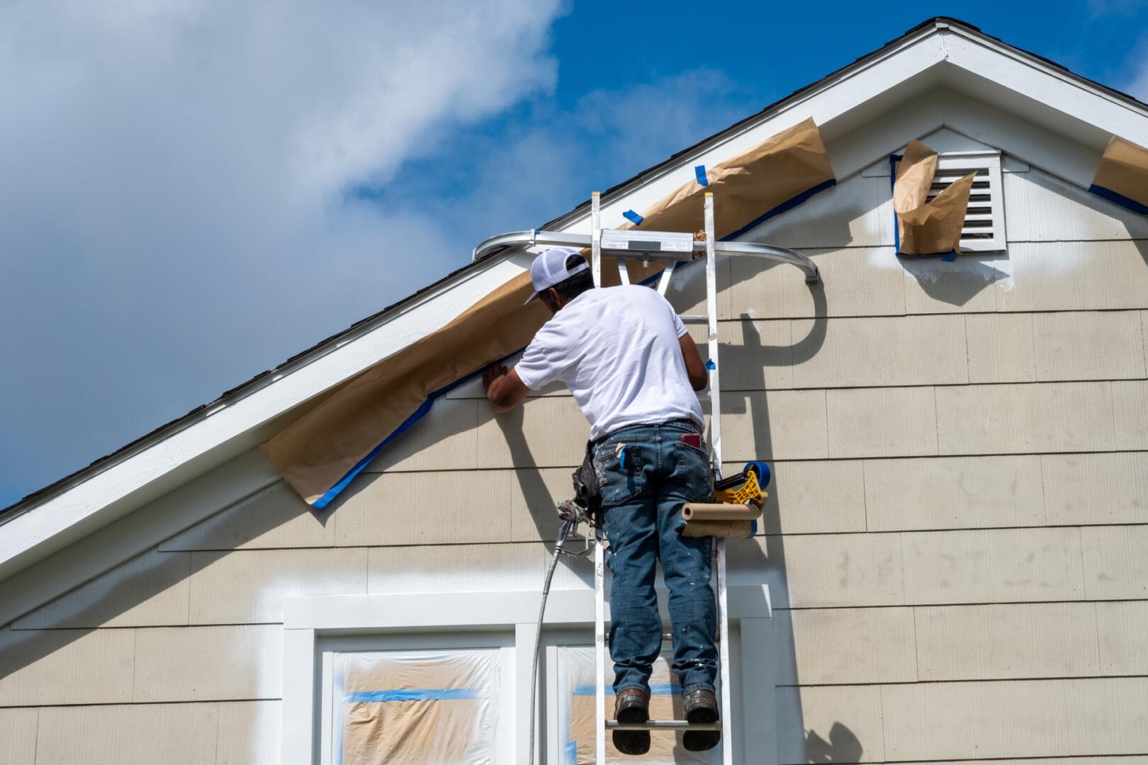 Painter on a ladder masking off trim at the peak of a house before spraying paint